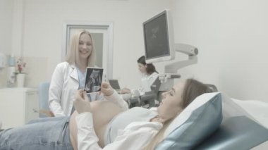 Kadın doktor ultrason çekerken, hamile kadın sınav arabasında yalan söylüyor, mutlu kocayla konuşuyor ve el ele tutuşuyor.