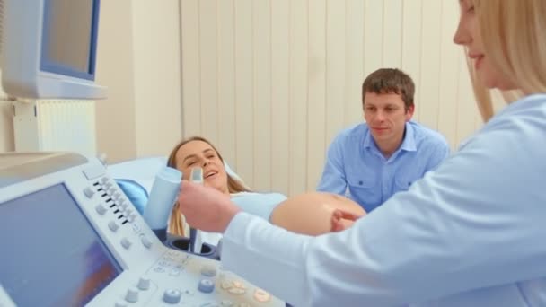 Schöne schwangere Frau, die auf einem Untersuchungsbus liegt und Ultraschallbilder des Babys mit ihrem Mann teilt, während Ärztin Rücksprache hält. — Stockvideo