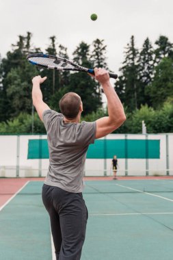 Tenis oynarken topu servis eden bir adamın arka görüntüsü. Erkekler güneşli bir günde dışarıda tenis oynuyorlar..
