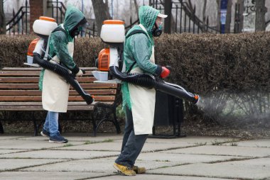 Koruyucu giysili iki adam karantina ve salgın sırasında bir şehir meydanını ya da sokağı dezenfekte ediyor. Antiviral Sıvı Spreyi. İhtiyati tedbirler.                               
