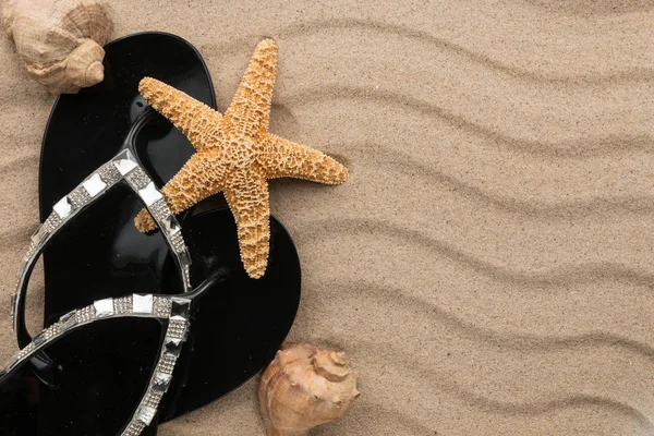 Chanclas negras con pedrería y concha, estrellas de mar en la arena ondulada . — Foto de Stock