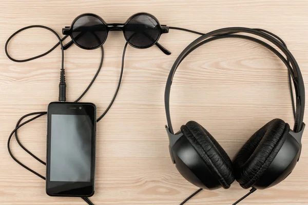Gafas de sol, teléfono y auriculares tumbados en una mesa de madera, con espacio para tu texto . — Foto de Stock