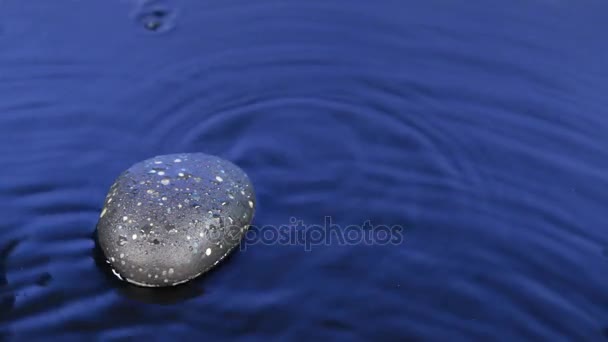 Il vento soffia su una goccia che cade nell'acqua blu e una pietra nera . — Video Stock