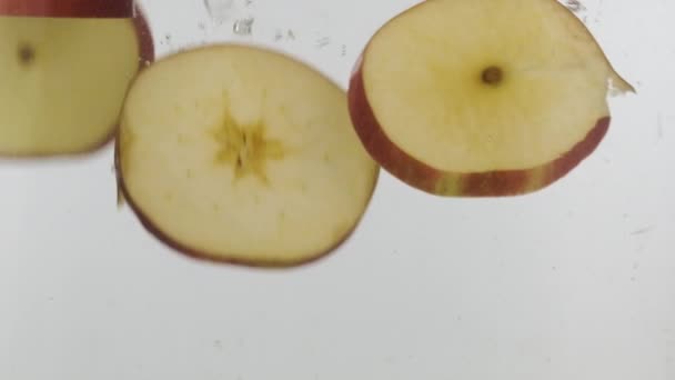 スローモーション。新鮮なスライスされた赤いリンゴは、白い背景にスプラッシュや泡で水に落ちます. — ストック動画
