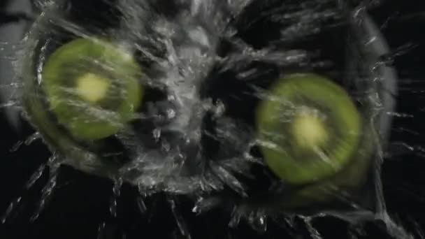 Sakta i backarna. Faller två skivor grön kiwi i vatten på svart bakgrund. Ovanifrån. — Stockvideo