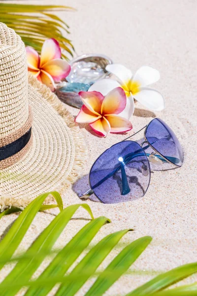 米色草帽与蓝色太阳镜 五颜六色的贝壳 弗兰吉帕尼花和绿棕榈叶上的沙 暑假的概念 垂直组成 — 图库照片