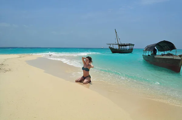 a young woman kneeling on a white sandy beach of Zanzibar in the Indian Ocean with its turquoise waters, two old vintage fishing boats, and cloudless sky on a sunny warm day