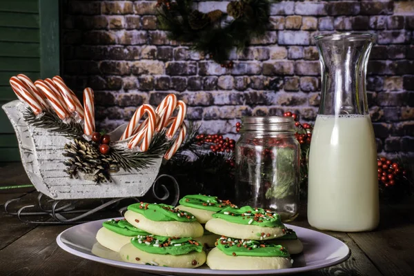 sugar cookies on white plate decorated with green icing and sprinkles on rustic wood table with candy canes and milk with Christmas wreath on brick wall background