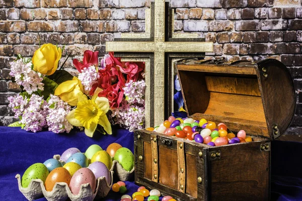 wood chest full or jelly beans next to tray of colored eggs on blue with spring flowers and wood cross and brick wall background