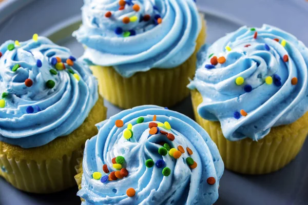close up of four yellow cupcakes with blue frosting and rainbow sprinkles on blue plate