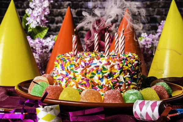 birthday cake with rainbow sprinkles on red plate surrounded with assorted gumdrops with smoking candles party hats party favors and spring flowers with brick wall background