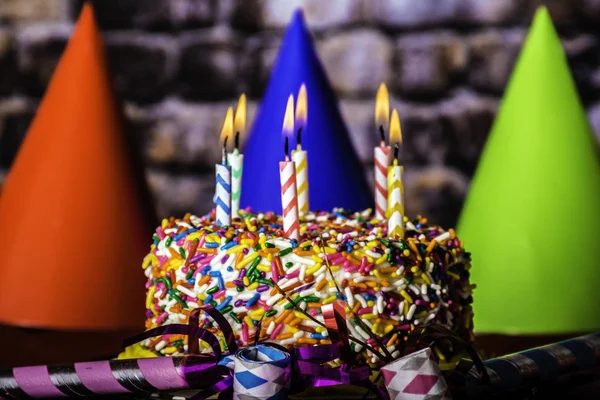 birthday cake with rainbow sprinkles surrounded by party favors and party hats with burning candles and brick wall background