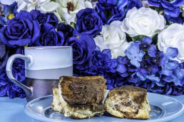 Mavi kahve fincanı mavi masanın üzerinde mavi ve beyaz bahar çiçekleri ile mavi tabağın üzerinde yapışkan çörekler.