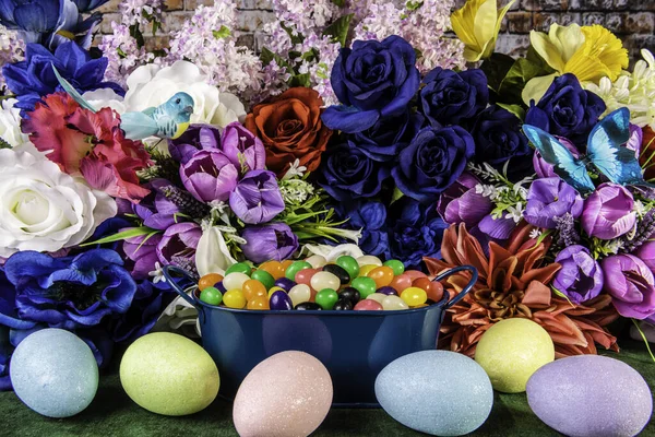 蓝色平底锅 盛满了果冻豆 周围环绕着五颜六色的彩蛋 有春天的花朵和砖墙背景 — 图库照片
