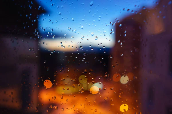 Wet window glass with rain drops in the evening. View on city with street lights in blur bokeh