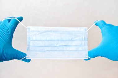 Tek kullanımlık mavi eldivenli bir adam gri arka planda tıbbi maske tutuyor. Acil tıbbi bakım.