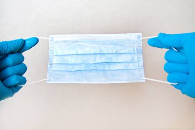 Tek kullanımlık mavi eldivenli bir adam gri arka planda tıbbi maske tutuyor. Acil tıbbi bakım.
