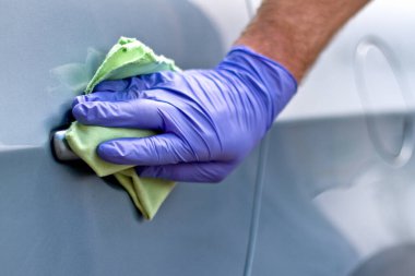 Erkek eli koruyucu eldivenlerle arabanın kapı kolunu antibakteriyel ya da temizlik solüsyonu kullanarak temizliyor..