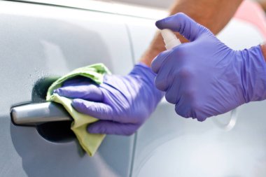 Erkek eli koruyucu eldivenlerle arabanın kapı kolunu antibakteriyel ya da temizlik solüsyonu kullanarak temizliyor..