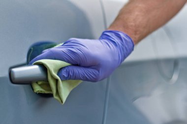 Erkek eli koruyucu eldivenlerle arabanın kapı kolunu antibakteriyel ya da temizlik solüsyonu kullanarak temizliyor..
