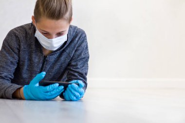 Çocuk evde karantina sırasında maske takmış telefonlar ve lastik eldivenlerle yerleri yalıyor..