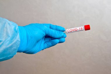 Üzerinde COVID-19 yazan gri arkaplandaki analizleri toplamak için elinde boş bir test tüpü bulunan mavi tek kullanımlık lastik eldivenleri teslim et..