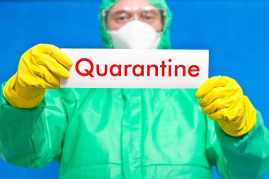 Üzerinde karantina yazılı beyaz bir levha tutan bir tıp çalışanı mavi arka planda duruyor..