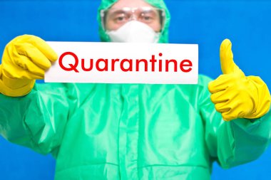 Üzerinde karantina yazılı beyaz bir levha tutan bir tıp çalışanı mavi arka planda duruyor..