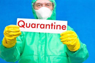 Üzerinde karantina yazılı beyaz bir levha tutan bir tıp çalışanı mavi arka planda duruyor..