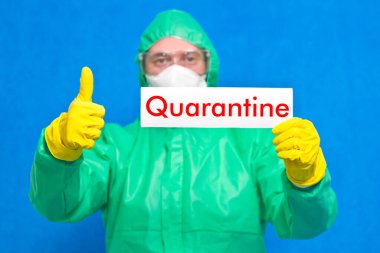 Üzerinde karantina yazılı beyaz bir levha tutan bir tıp çalışanı mavi arka planda duruyor..