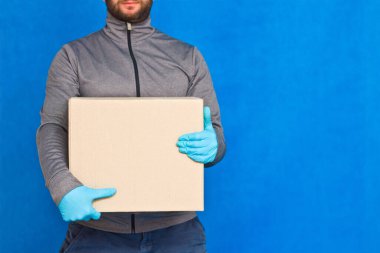 Kurye, mavi arka planda kahverengi bir kutu tutuyor. Yüz maskesi takmış lastik eldivenlerle. Kopya alanı olan karton bir paket taşıyan bir adamın ayrıntıları