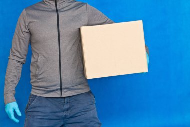 Kurye, mavi arka planda kahverengi bir kutu tutuyor. Yüz maskesi takmış lastik eldivenlerle. Kopya alanı olan karton bir paket taşıyan bir adamın ayrıntıları