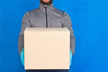 Kurye, mavi arka planda kahverengi bir kutu tutuyor. Yüz maskesi takmış lastik eldivenlerle. Kopya alanı olan karton bir paket taşıyan bir adamın ayrıntıları