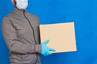 Kurye, mavi arka planda kahverengi bir kutu tutuyor. Yüz maskesi takmış lastik eldivenlerle. Kopya alanı olan karton bir paket taşıyan bir adamın ayrıntıları