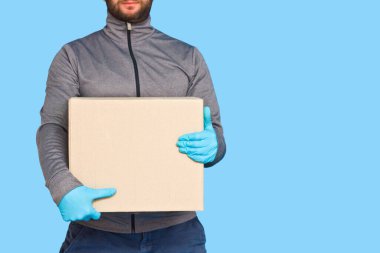 Kurye, maske takan lastik eldivenlerle mavi arka plandaki izolasyonda kahverengi kutuyu tutuyor. Kopya alanı olan karton bir paket taşıyan bir adamın ayrıntıları