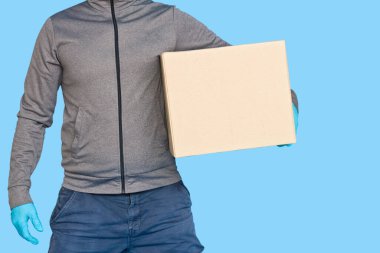 Kurye, maske takan lastik eldivenlerle mavi arka plandaki izolasyonda kahverengi kutuyu tutuyor. Kopya alanı olan karton bir paket taşıyan bir adamın ayrıntıları