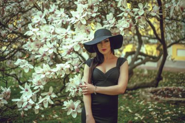 Manolya çiçek bir arka plan üzerinde siyah elbiseli siyah saçlı güzel kız. Arka plan bulanıklık. Açık havada çekim. Moda ve güzellik kavramı. Güzel kız ve Manolya çiçek. Sping ağaç çiçek.