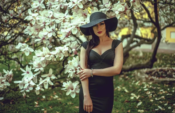 Belle fille aux cheveux noirs dans une robe noire sur un fond de fleur de magnolia. Fond flou. Tirer à l'extérieur. Le concept de mode et de beauté. Belle fille et fleurs de magnolia. Sping fleurs d'arbre . — Photo