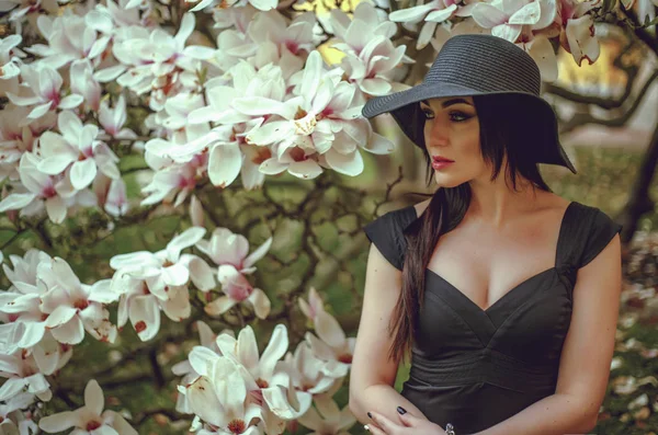 Hermosa chica con el pelo negro en un vestido negro sobre un fondo de una flor de magnolia. Un fondo borroso. Disparando al aire libre. El concepto de moda y belleza. Hermosa chica y magnolia flores. Sping flores de árbol . — Foto de Stock