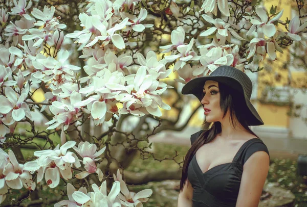 Hermosa chica con el pelo negro en un vestido negro sobre un fondo de una flor de magnolia. Un fondo borroso. Disparando al aire libre. El concepto de moda y belleza. Hermosa chica y magnolia flores. Sping flores de árbol . — Foto de Stock