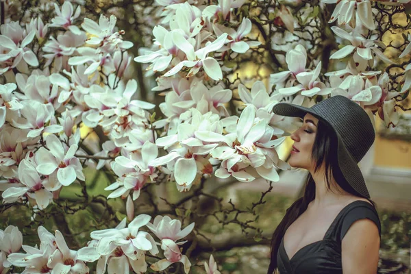 Hermosa chica con el pelo negro en un vestido negro sobre un fondo de una flor de magnolia. Un fondo borroso. Disparando al aire libre. El concepto de moda y belleza. Hermosa chica y magnolia flores. Sping flores de árbol . — Foto de Stock
