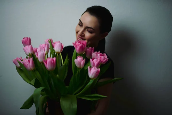 Young beautiful happy girl holding spring bouquet of tulips, posing on white background.  Portrait of attractive young woman with tulips is standing in light room and smiling. Happy international women's day! Celebrating 8th of March.