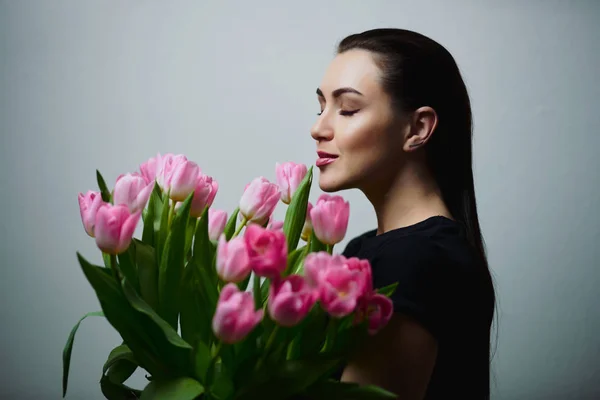 Young beautiful happy girl holding spring bouquet of tulips, posing on white background.  Portrait of attractive young woman with tulips is standing in light room and smiling. Happy international women\'s day! Celebrating 8th of March.