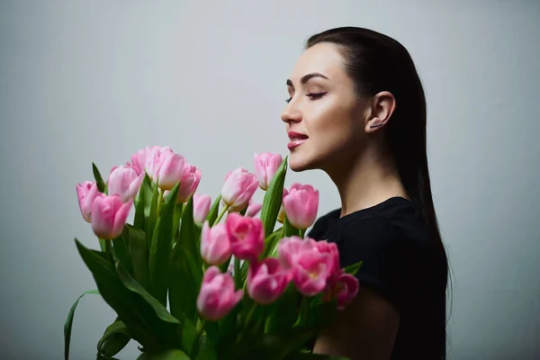 Young beautiful happy girl holding spring bouquet of tulips, posing on white background.  Portrait of attractive young woman with tulips is standing in light room and smiling. Happy international women\'s day! Celebrating 8th of March.