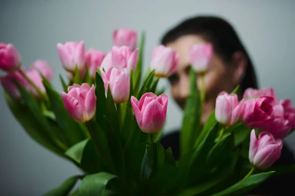 Young beautiful happy girl holding spring bouquet of tulips, posing on white background.  Portrait of attractive young woman with tulips is standing in light room and smiling. Happy international women's day! Celebrating 8th of March.