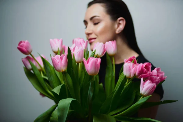 Young beautiful happy girl holding spring bouquet of tulips, posing on white background.  Portrait of attractive young woman with tulips is standing in light room and smiling. Happy international women\'s day! Celebrating 8th of March.