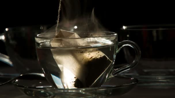 Sachet de thé dans une tasse transparente. Teabag tombe dans une tasse transparente avec de l'eau chaude sur un fond sombre. L'eau est peinte dans la couleur du thé. Tournage à un rythme de 240 images par seconde .
