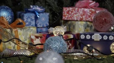 Hediyeler ve Noel süsleri. Kamera, parıltılı fiyonklarla süslenmiş hediye kutuları, Noel ağacı süslemeleri ve ışıltılı çelenklerle yavaşça hareket ediyor.