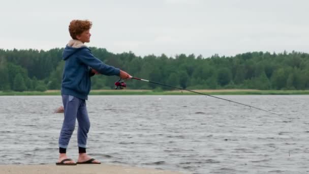 桟橋で釣りをする少年 水平線の湖と森の背景に回転を使用してスピナーをスローします — ストック動画