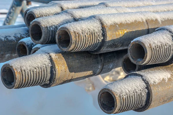 Drill pipes are located horizontally on the rack. In the foreground, a threaded joint of pipes. Rust is visible on the pipes. On top of the pipes is snow.